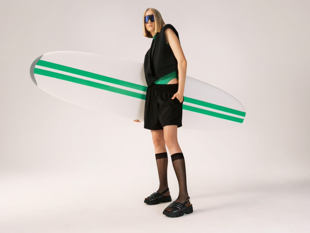 Modelo en total look negro con sandalias negras, sosteniendo una tabla de surf - Campaña verano 2023